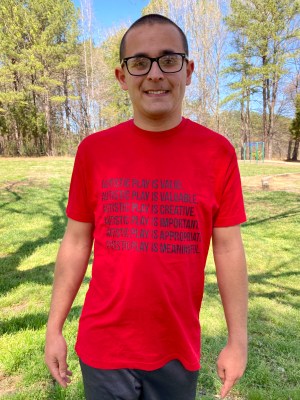 Nicholas Hemachandra in a bright red shirt that says; "Autistic play is valid. Autistic play is valuable. Autistic play is creative. Autistic play is important. Autistic play is appropriate. Autistic play is meaningful."